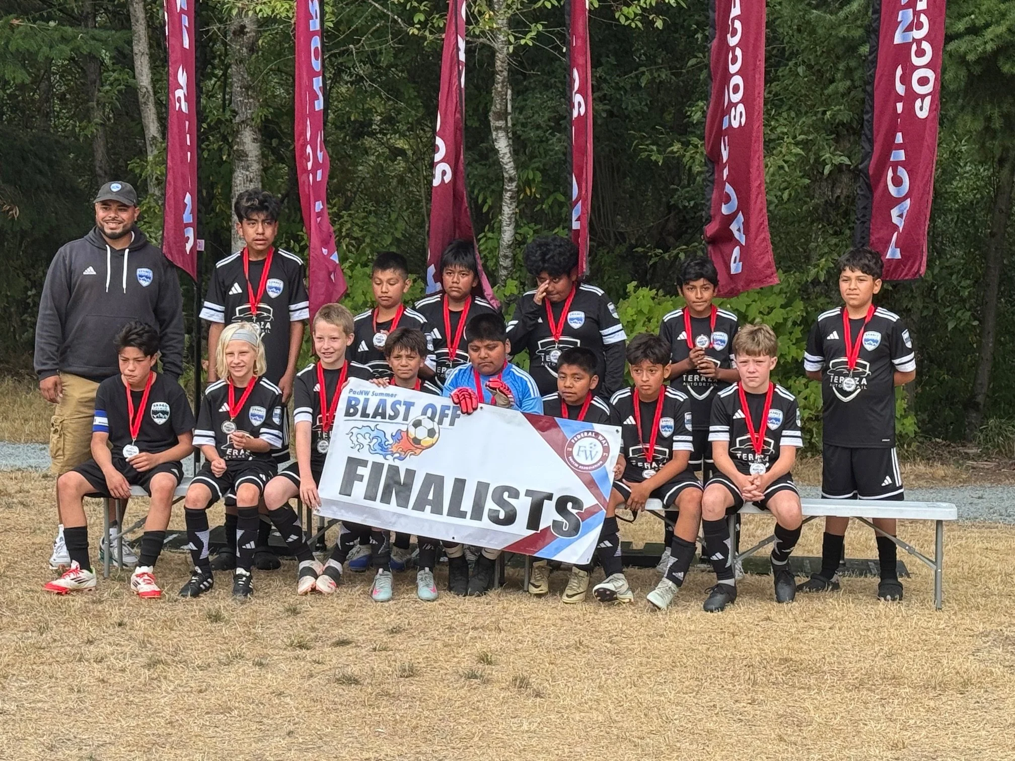 A youth soccer team, mostly boys and one girl, posing outdoors with their coach. They're wearing black uniforms with white accents, medals around their necks, and holding a banner that says 'Blast Off Finalists.' Some players have soccer cleats, and the scene includes a backdrop of trees and red flags.