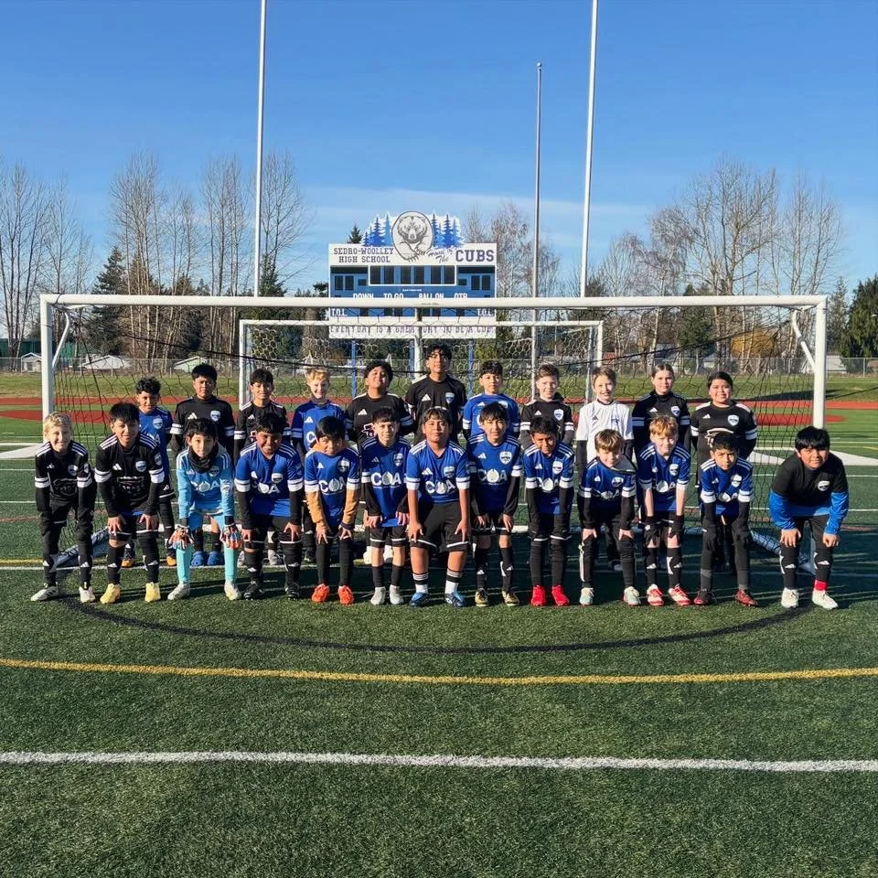 Youth soccer team posing for a group photo on a field with goalposts in the background, wearing black and blue jerseys.