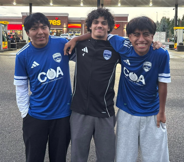 Three young boys standing together outdoors in front of a gas station, smiling, with two of them wearing blue soccer jerseys and the middle boy in a black jacket.