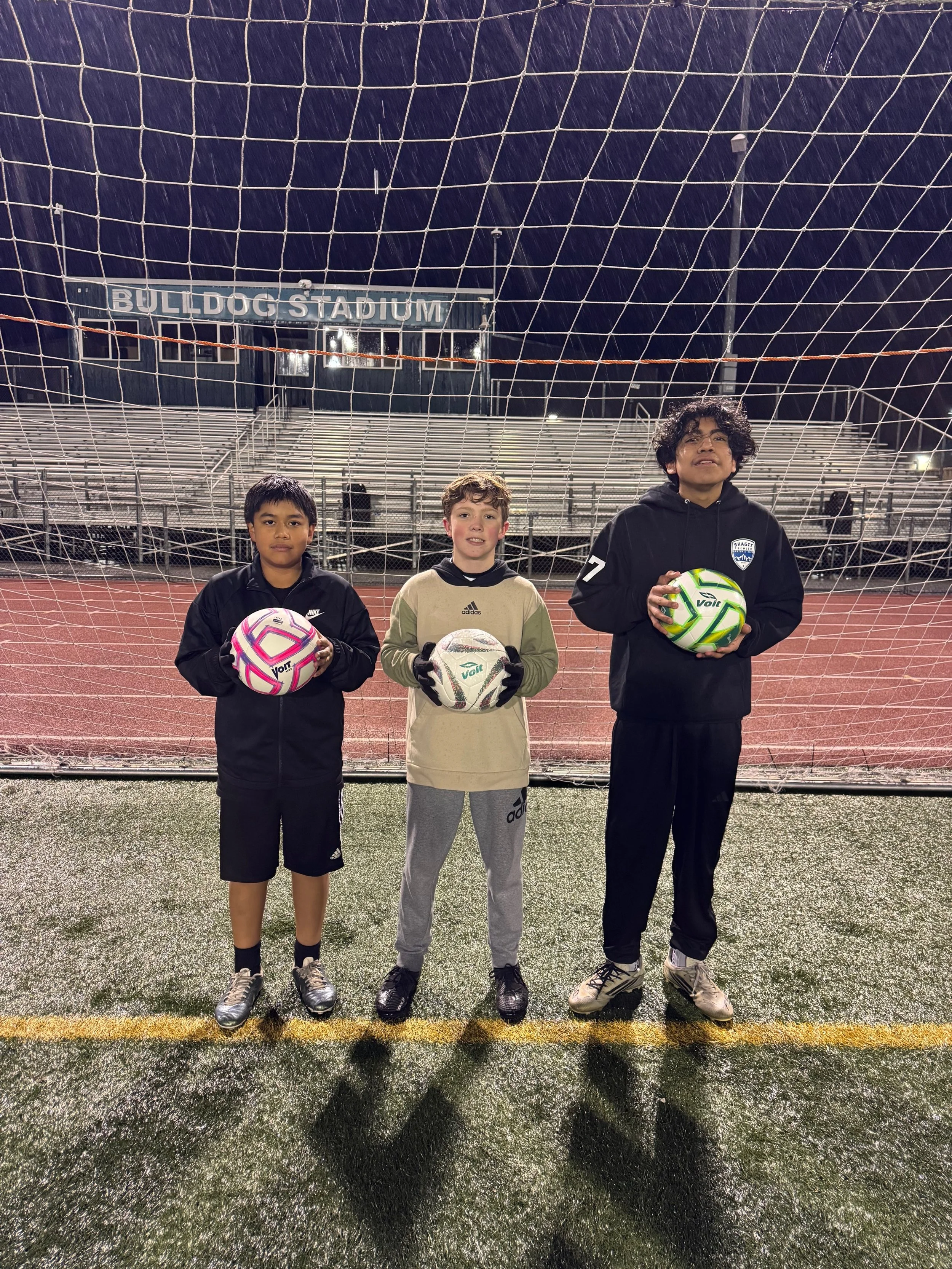 Three young boys standing on a soccer field at night, holding soccer balls, in front of a goal with a sign that says 'Bulldog Stadium'.