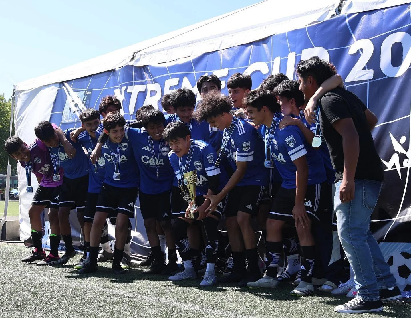 A group of young male soccer players in blue uniforms with medals around their necks are celebrating together on a field after winning a trophy, overseen by a person in black and blue jeans.