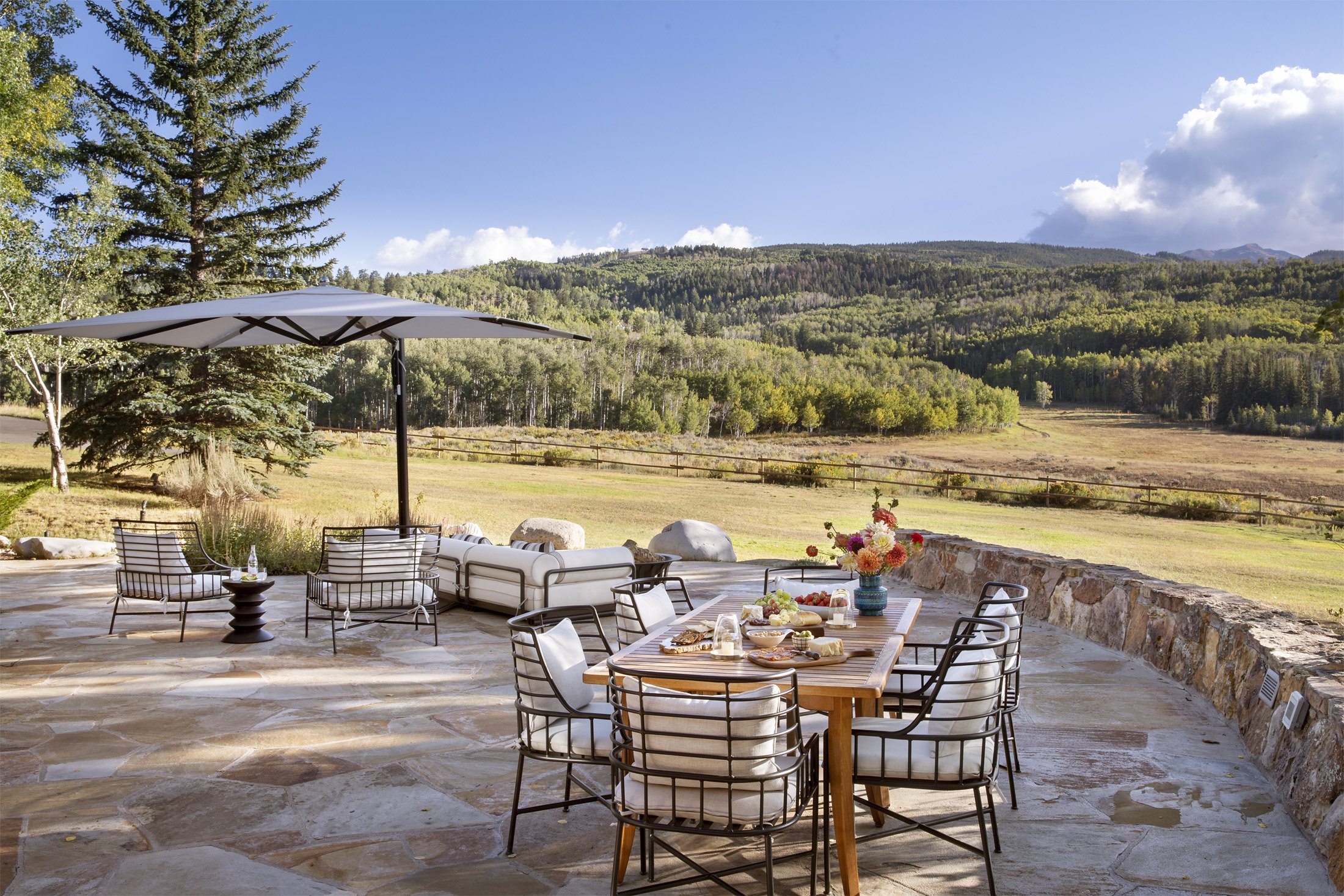 Outdoor patio with a dining table set with a floral centerpiece and food, surrounded by chairs and lounge seating, overlooking a grassy landscape with trees and mountains in the background under a partly cloudy sky.