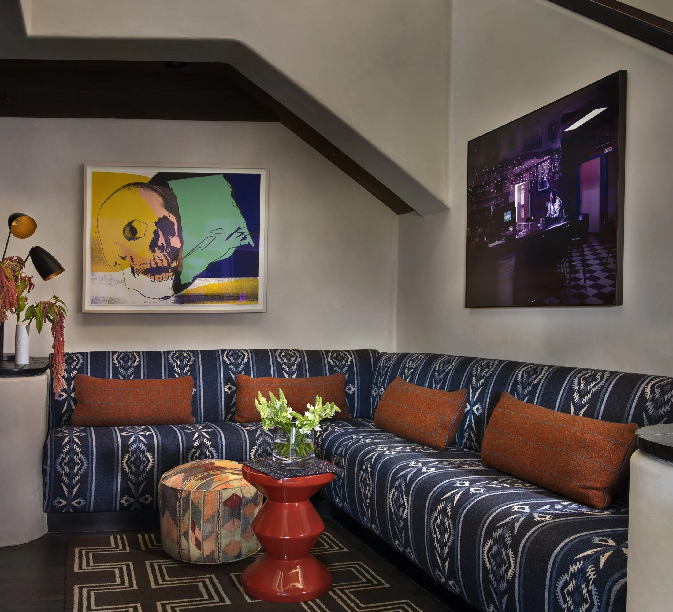 Living room with a patterned blue sofa, orange pillows, a round patterned ottoman, a red side table with a glass vase of green plants, artwork on the wall including a colorful skull and a neon-lit scene, and a black and white patterned rug.