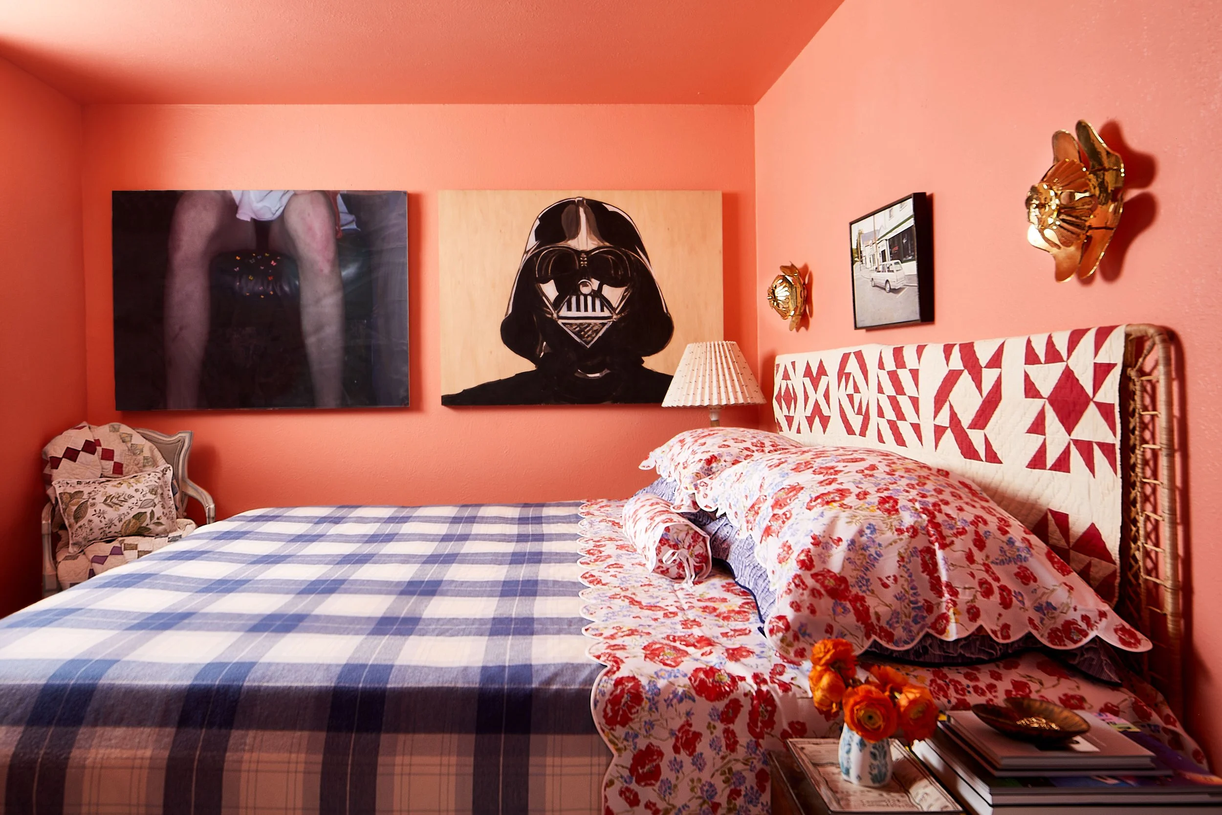 Bedroom with pink walls, decorated with artwork including a humorous portrait of Darth Vader and a photograph of a person with visible legs. The bed has a patterned red and white headboard, floral and checked bedding, and a nightstand with a vase of 