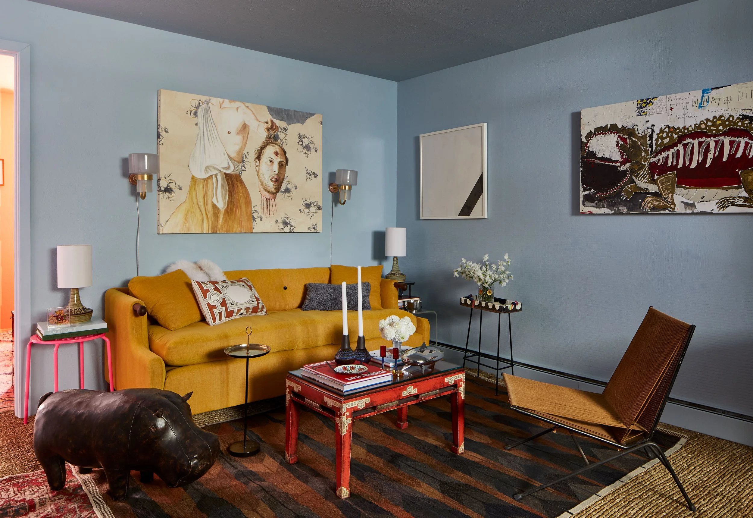 Living room with yellow sofa, abstract art on light blue walls, and mid-century modern furniture, including a brown chair and colorful decor items.