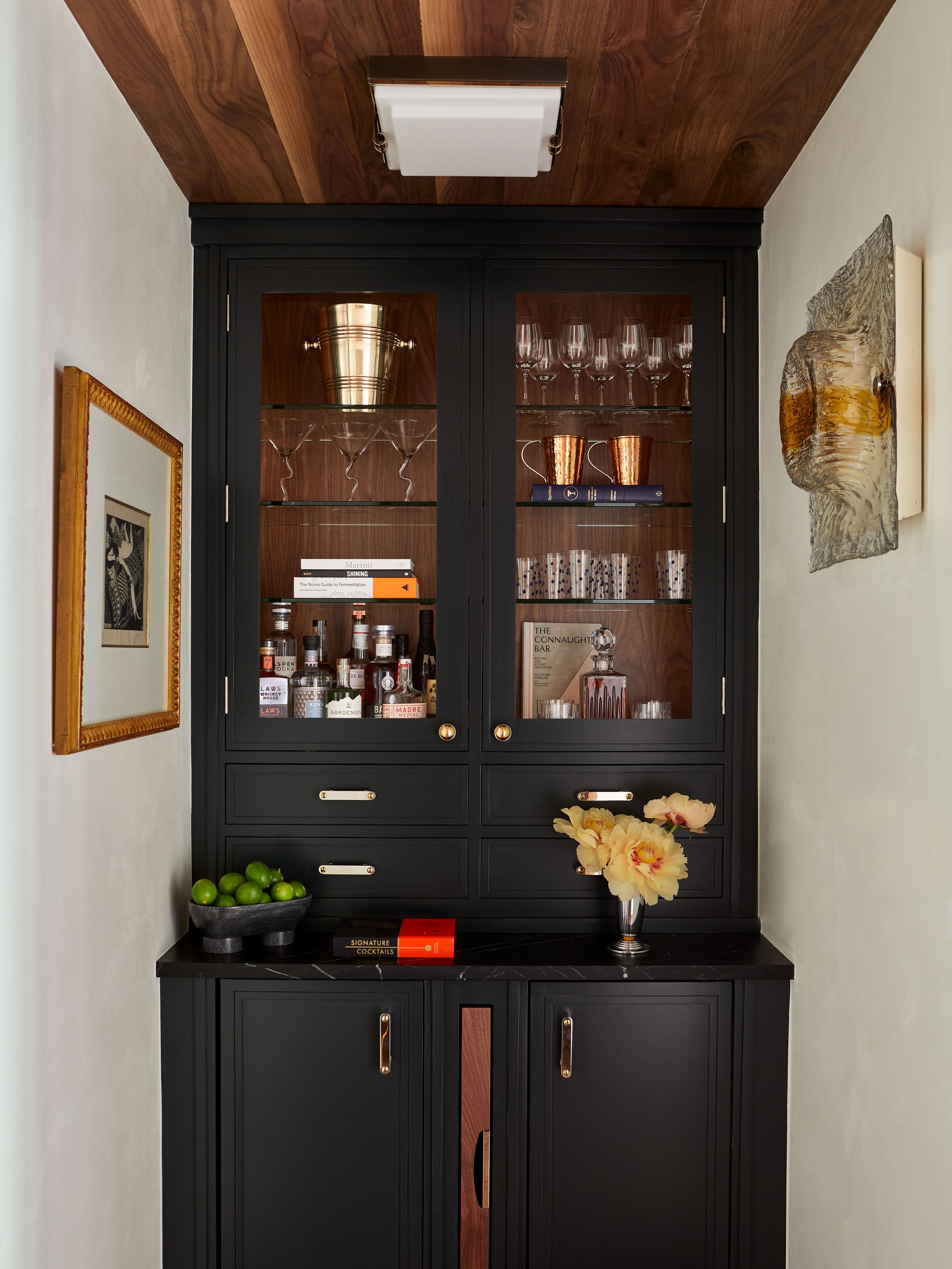A black wooden bar cabinet with glass-paneled doors showcasing glassware, bottles, and books, topped with a black marble surface with a bowl of limes and a floral arrangement, and adorned with wall art and a ceiling light.