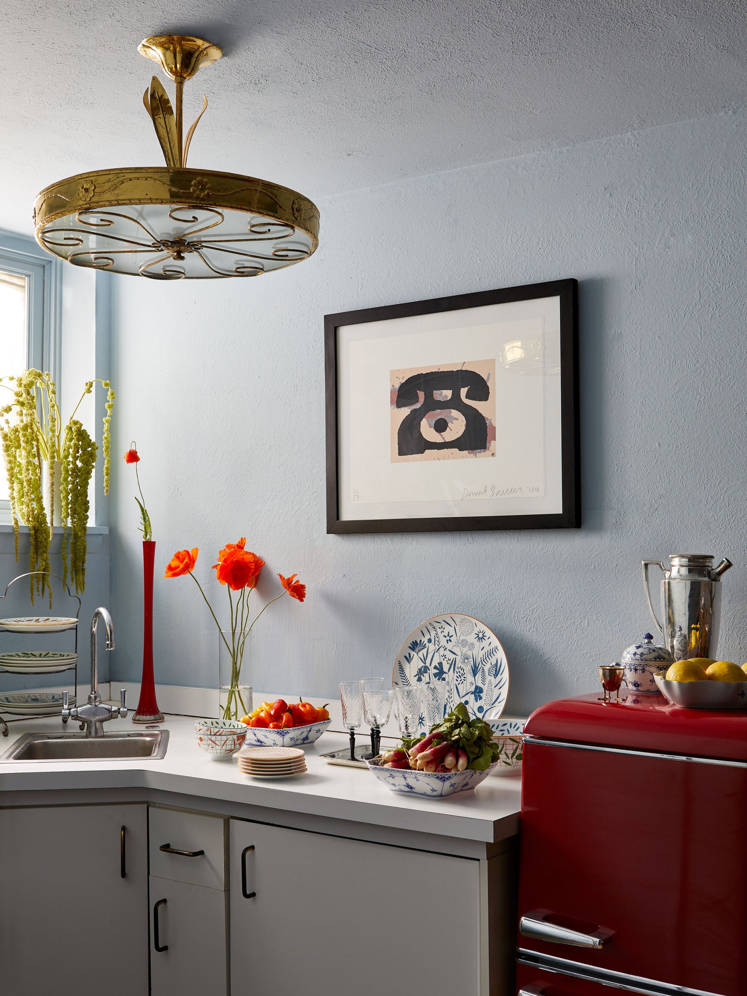A kitchen with a white countertop, a red refrigerator, and decorative items including flowers, dishes, and a framed piece of artwork on a light blue wall.