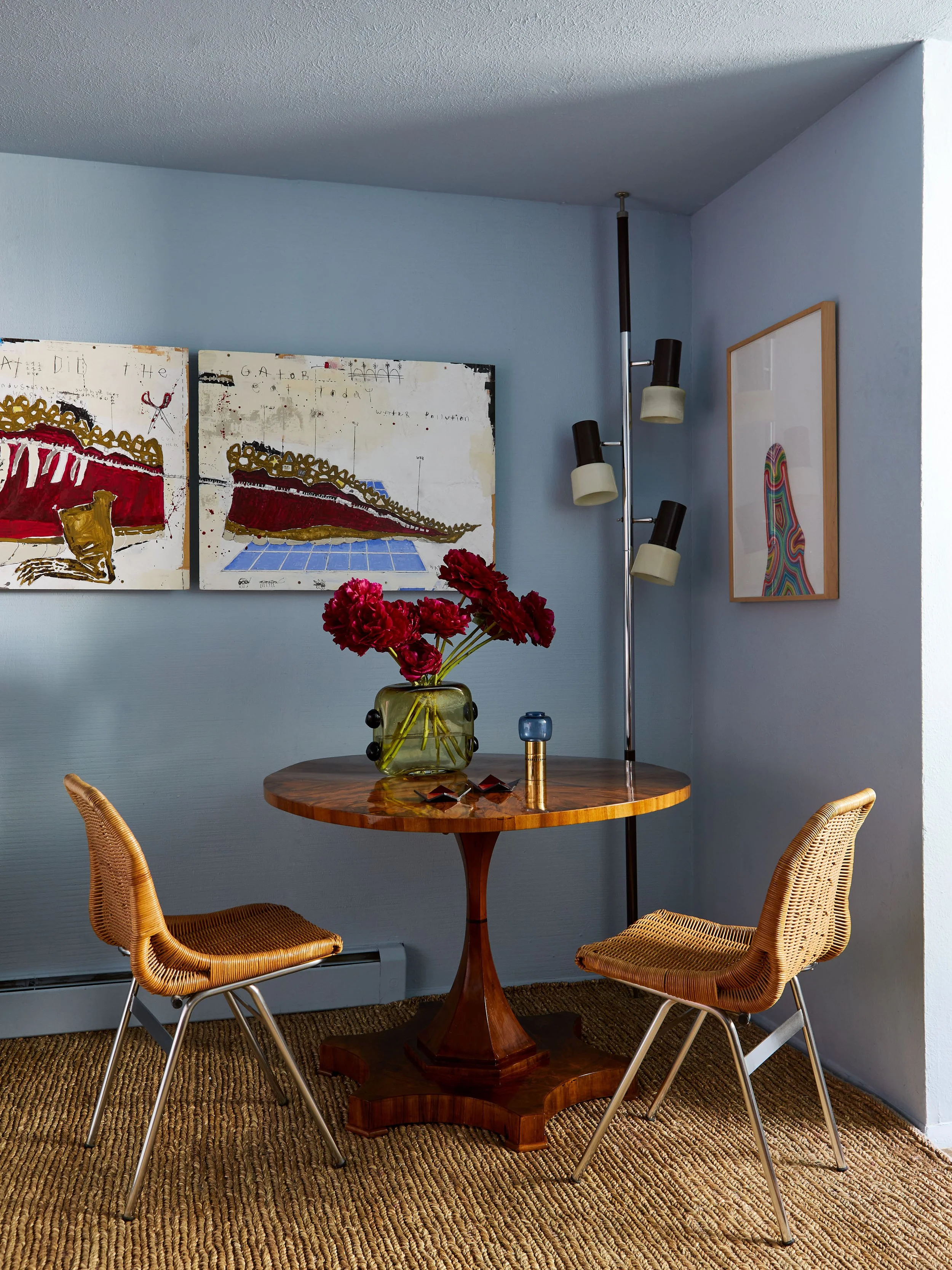 A small wooden round table with a glass vase of red flowers on top, two woven rattan chairs, a floor lamp with three adjustable black and white lamps, abstract wall art, and a textured brown rug on a light blue wall background.