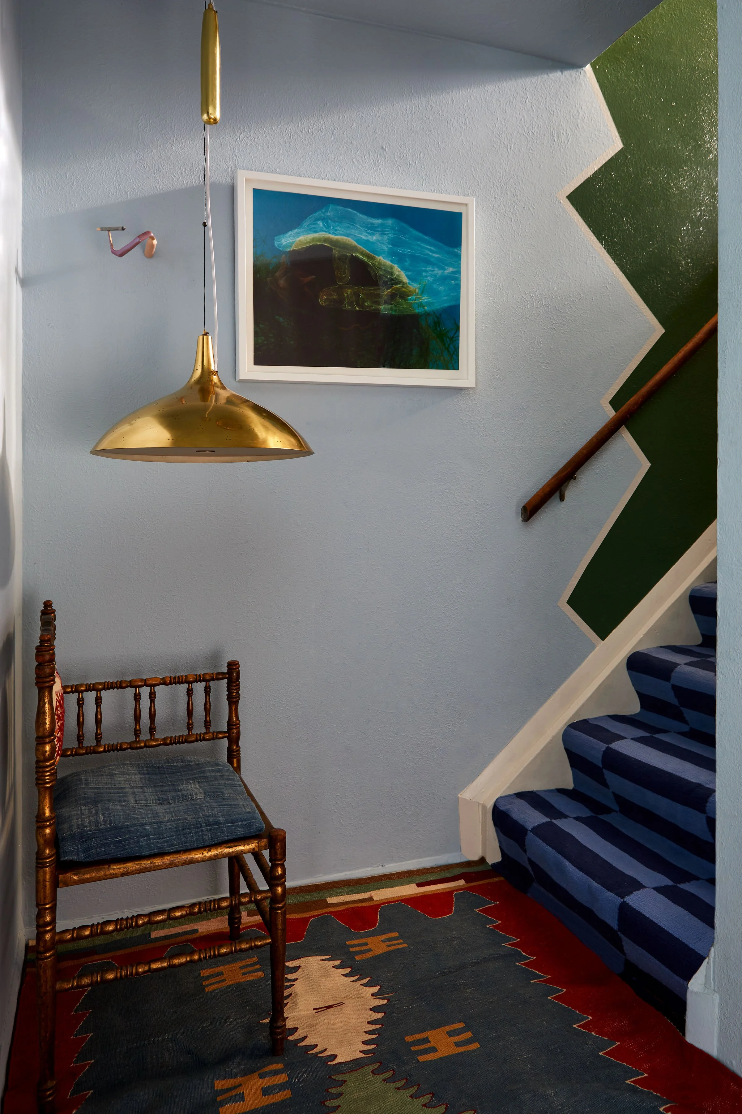 A staircase with a green zigzag wall, a blue patterned carpet, a wooden chair with a plaid cushion, a hanging gold lamp, and a framed underwater landscape photograph on the wall.