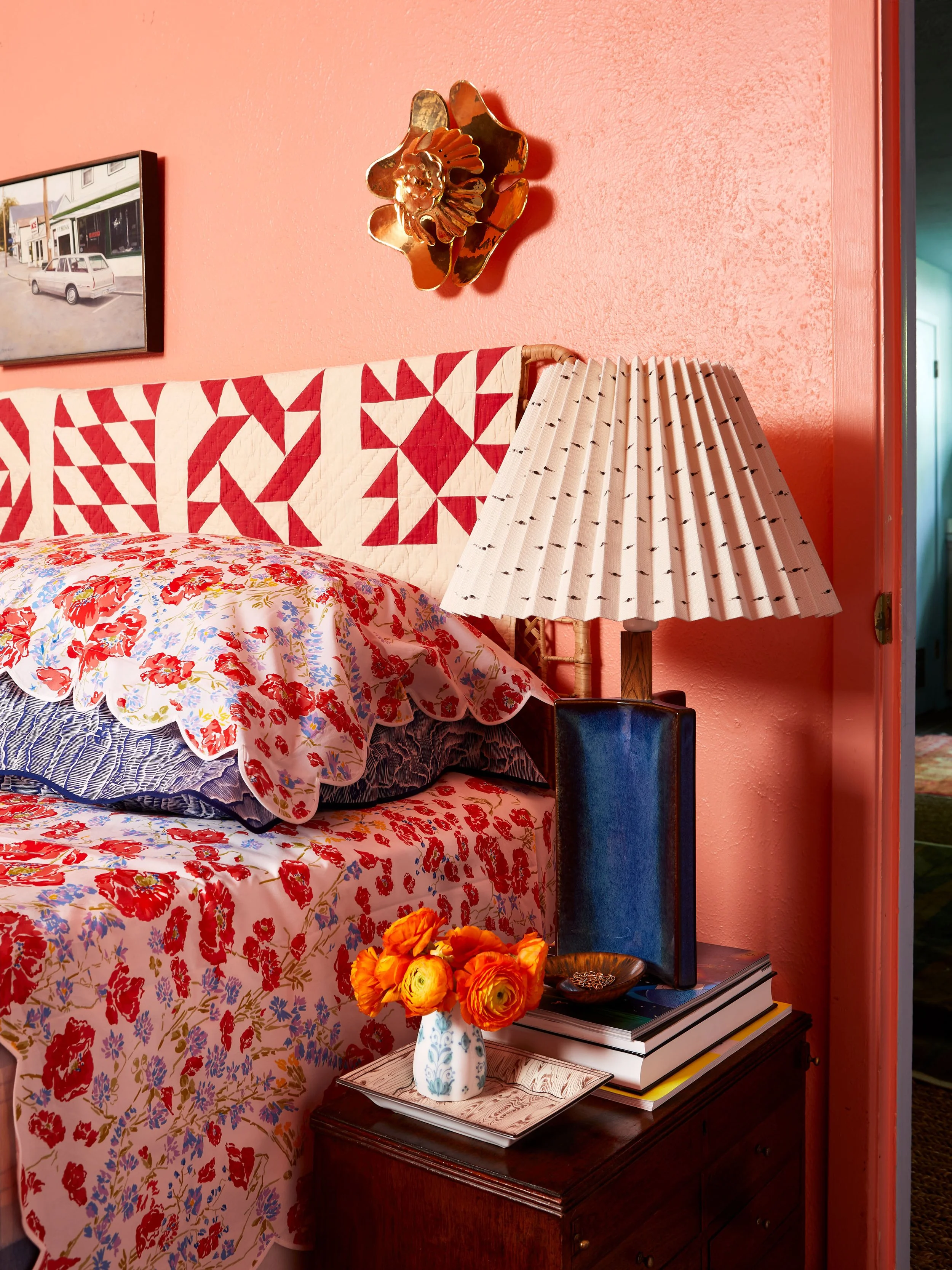 A bedroom corner with a floral-patterned bedspread, pillows, a bedside table with a vase of orange flowers, books, and a lamp with a pleated shade. The wall is painted pink with a framed picture and a gold decorative wall piece.