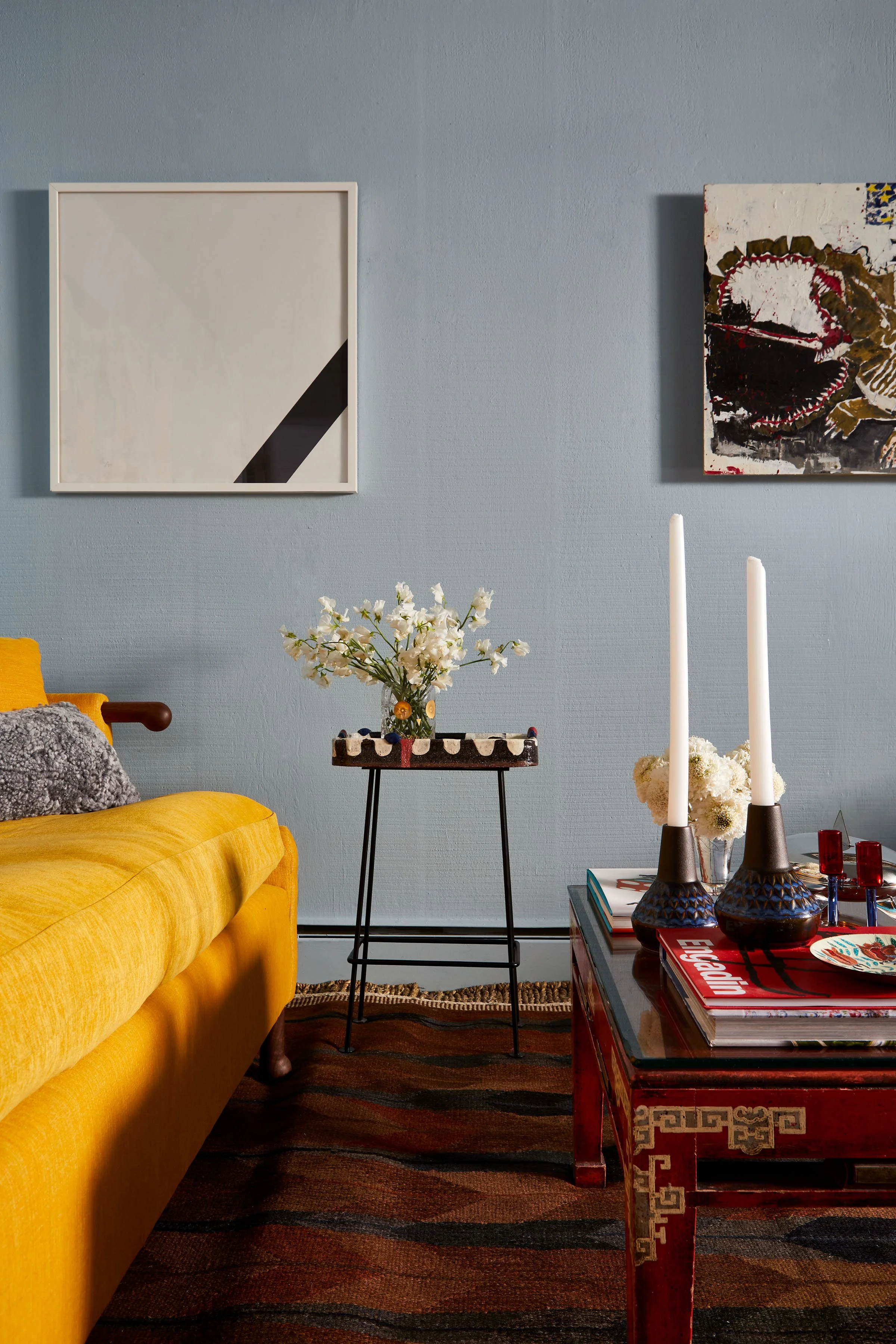 Living room with a yellow sofa, a small black side table with white flowers, a red coffee table with candles and magazines, and wall art including an abstract painting and a small square piece, against a blue wall.