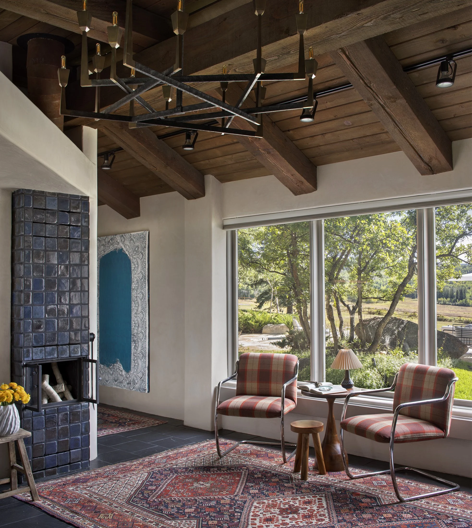 Living room with large windows showing outdoor trees and landscape, two plaid armchairs, a small round table with a lamp, a patterned rug, a wood ceiling with beams, and a modern chandelier.