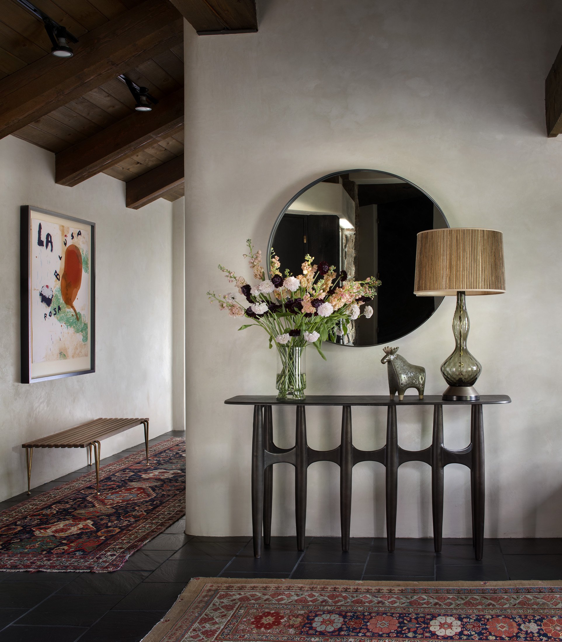 A hallway with a dark wooden ceiling, a decorative rug on black tiled floor, and a white textured wall. A black console table holds a glass vase with pink and dark purple flowers, a decorative metal animal figure, and a large glass table lamp with a 
