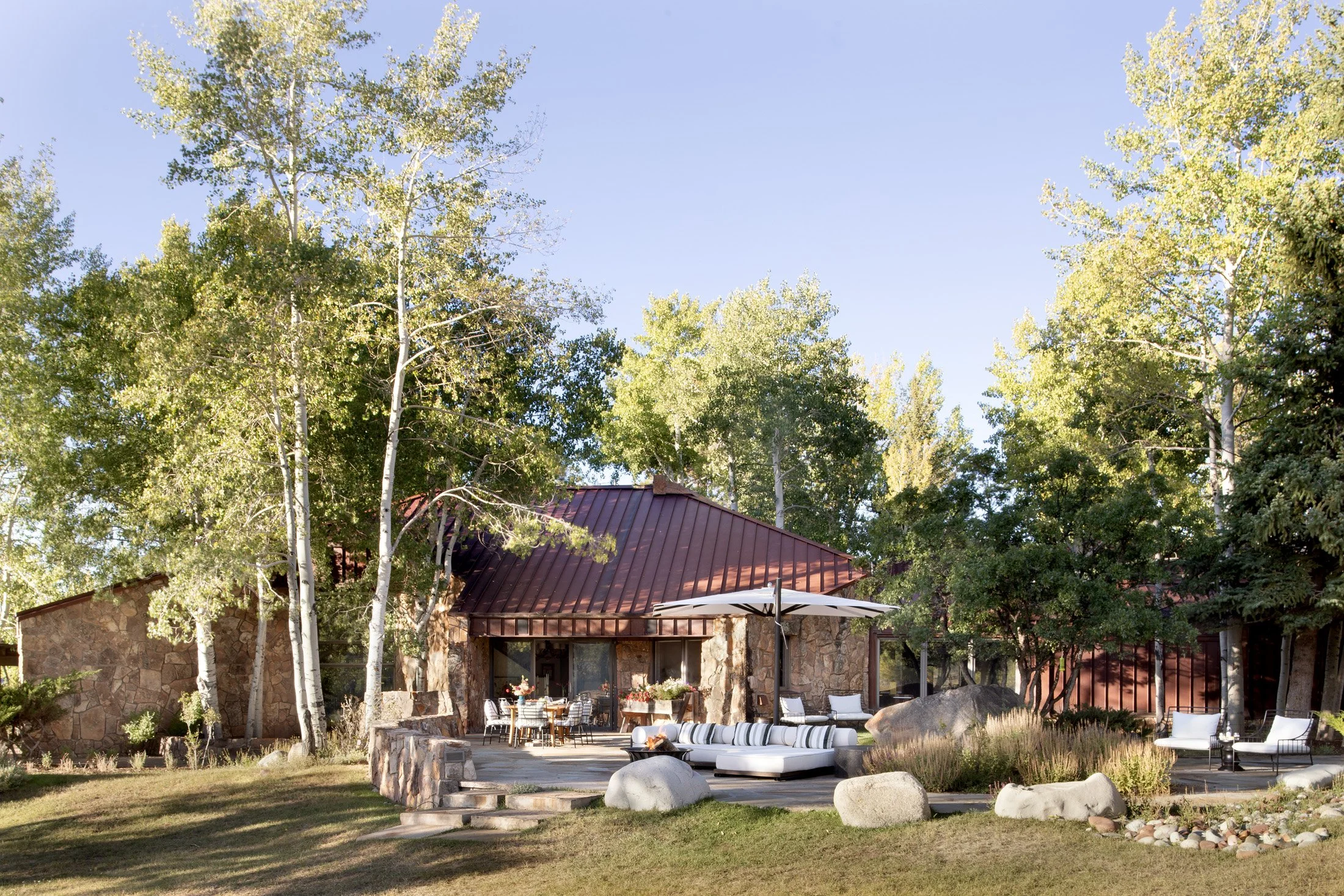 A house with a red metal roof surrounded by trees, with an outdoor seating area including a curved white sofa, chairs, tables, and an umbrella, set on a stone patio.