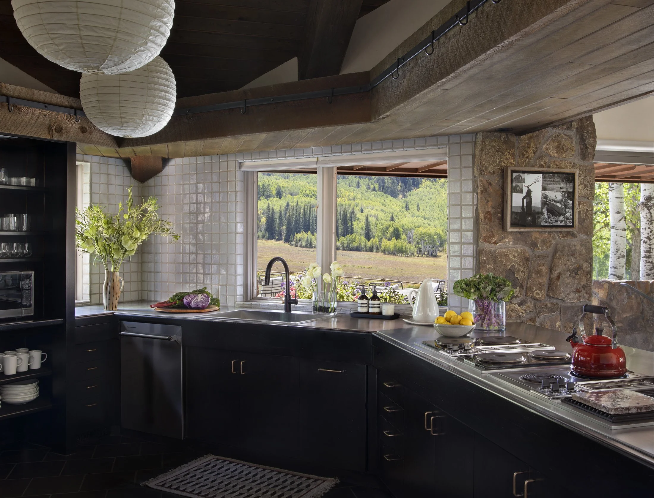 Modern kitchen with black cabinets, a window overlooking a green landscape, and decorative items including flowers, lemons, and a red kettle.