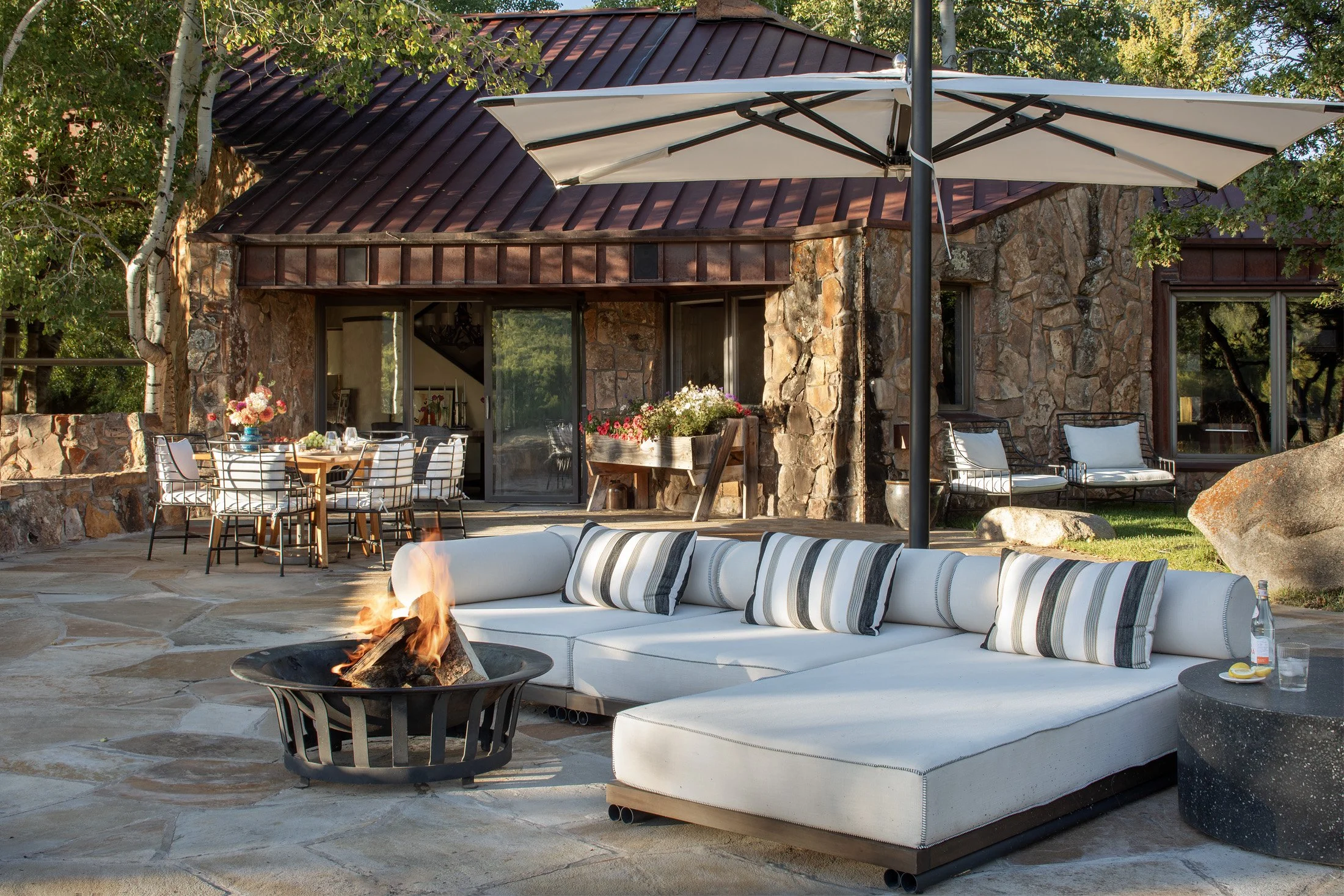 Outdoor patio with a modern sectional sofa around a fire pit, a large umbrella, a wooden dining table with chairs, and a stone house in the background.