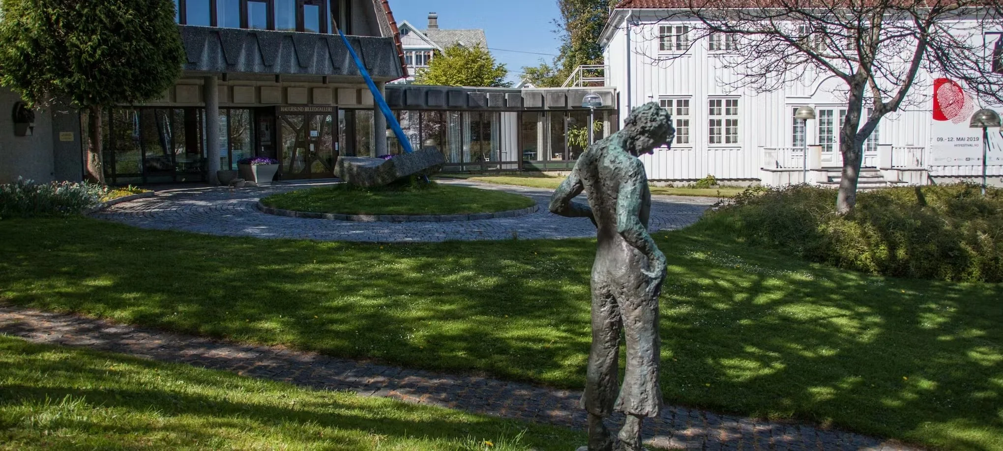 Skulptur av en menneskefigur i en park med gress, trær og bygninger i bakgrunnen.