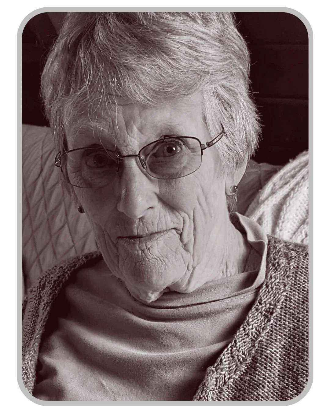 Close-up black and white portrait of an elderly woman with glasses, short gray hair, and earrings, sitting on a textured pillow or blanket.