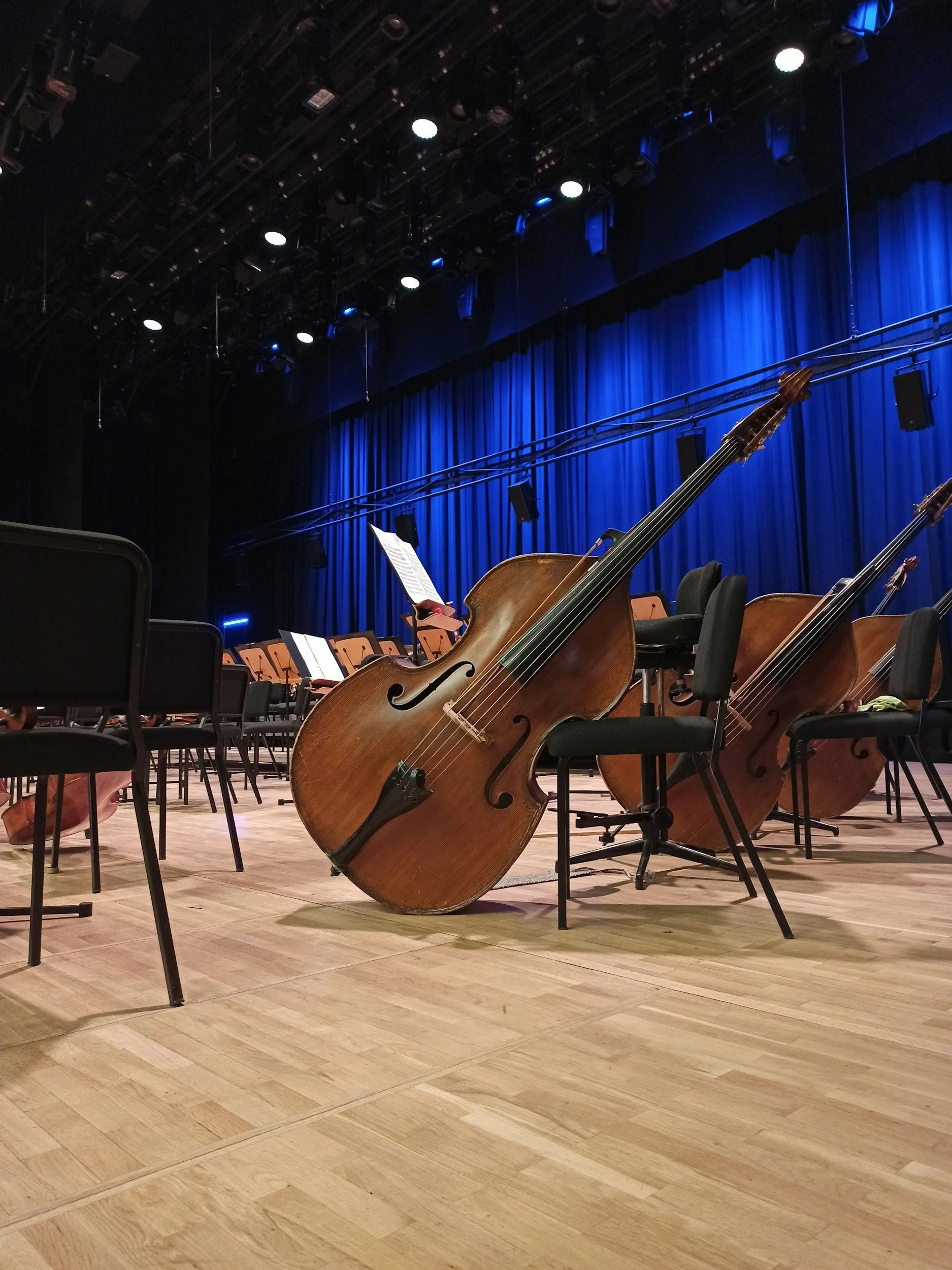 An empty concert hall with a double bass leaning against a chair, surrounded by other chairs and music stands, ready for a performance.