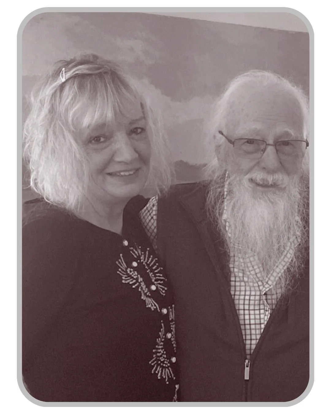 Black and white photo of a smiling woman with curly blonde hair and a man with glasses and a long white beard, both looking at the camera, with a cloudy sky in the background.