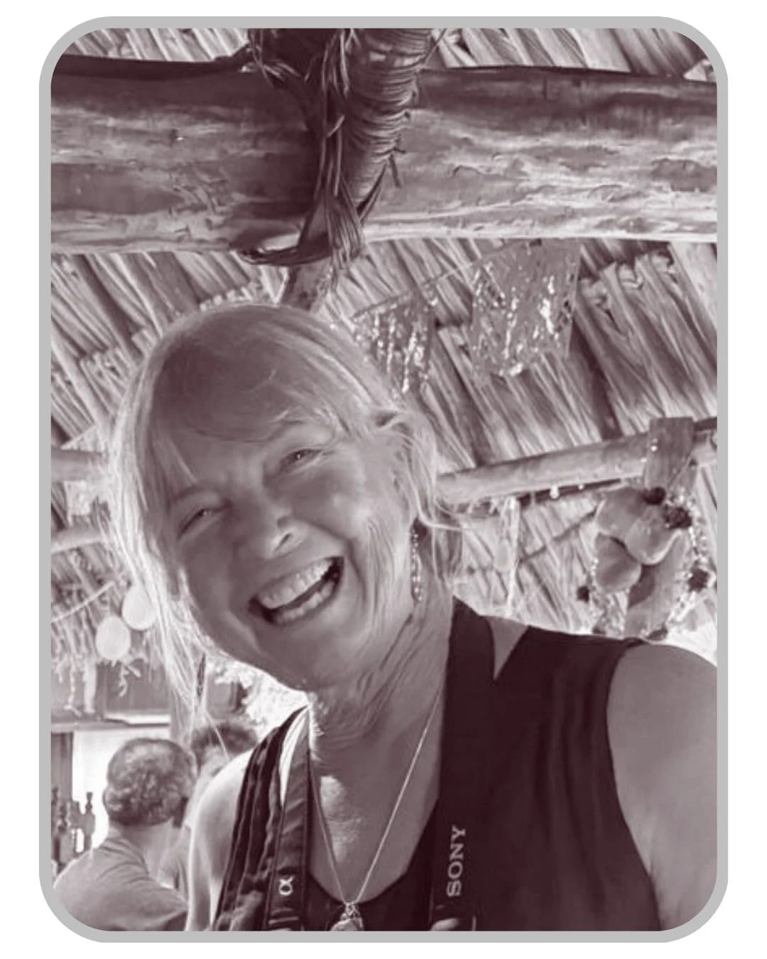 A woman with short hair and earrings smiling joyfully at the camera under a thatched roof with wooden beams.