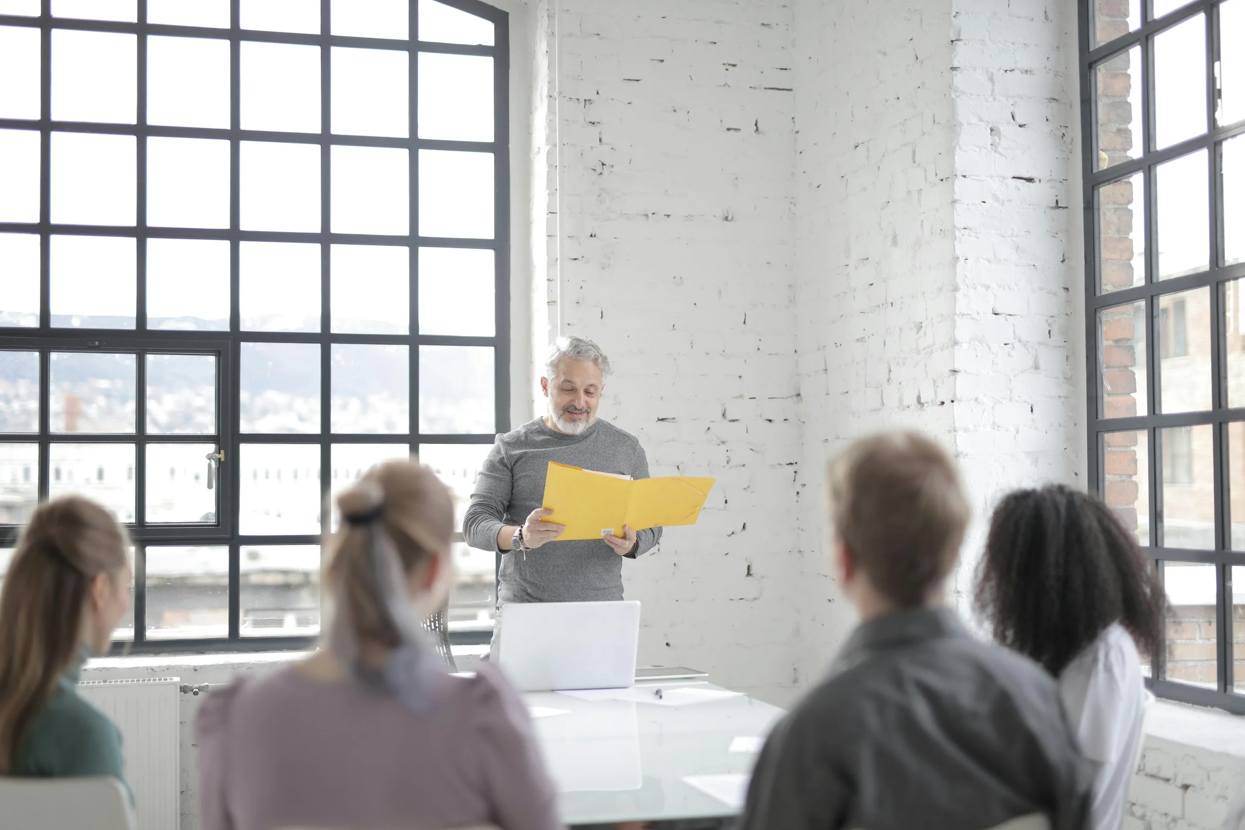 A man standing in front of a white brick wall, holding a yellow folder, giving a presentation to a group of four people seated at a table in a bright room with large windows.