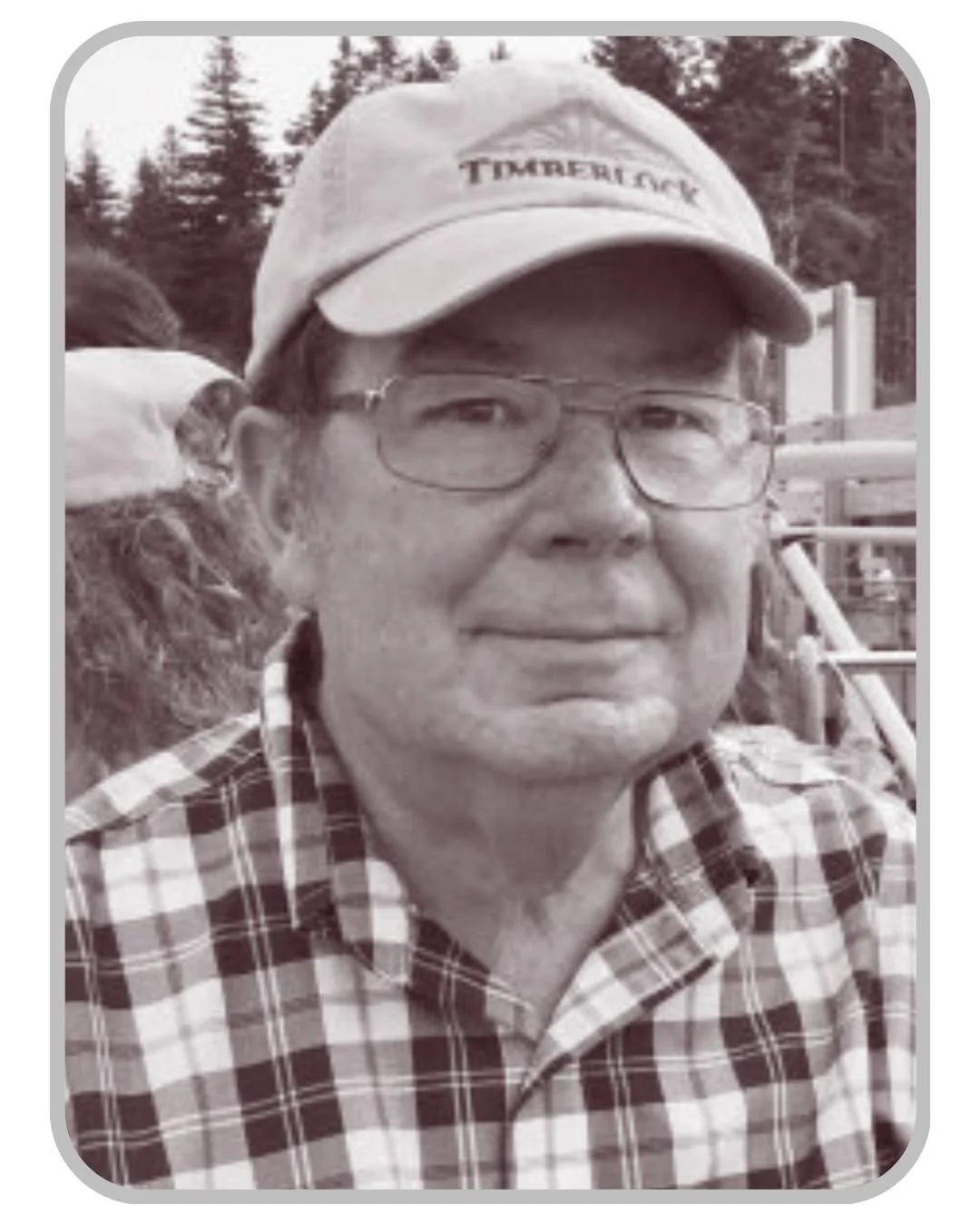 A man wearing glasses, a baseball cap, and a plaid shirt, smiling outdoors with trees in the background.
