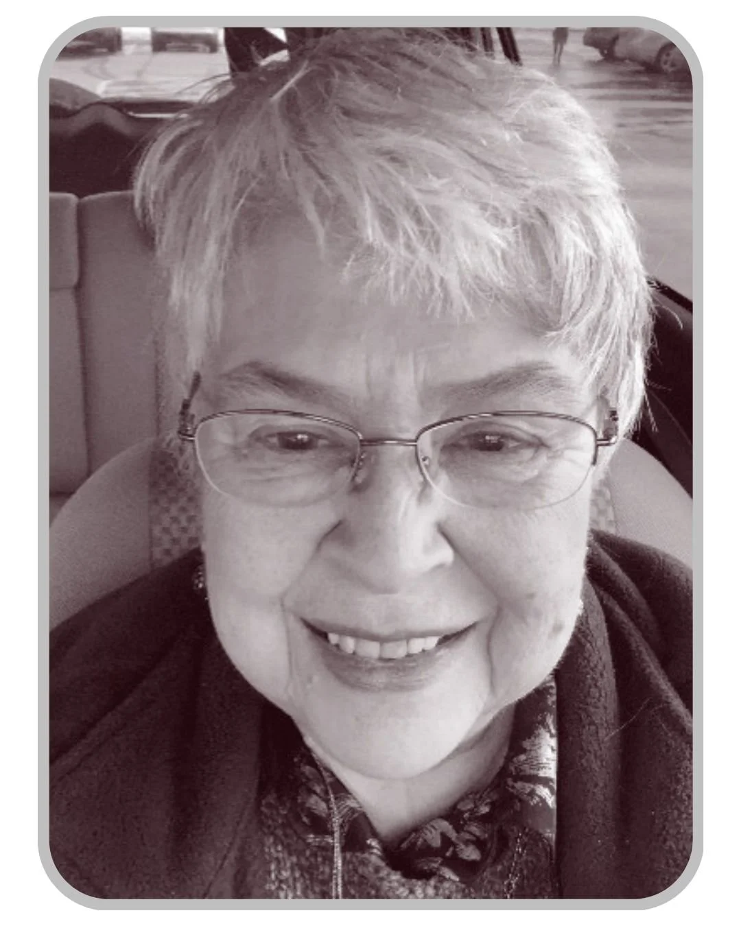 A close-up black and white photo of an elderly woman with short light-colored hair, glasses, smiling, sitting in a car.