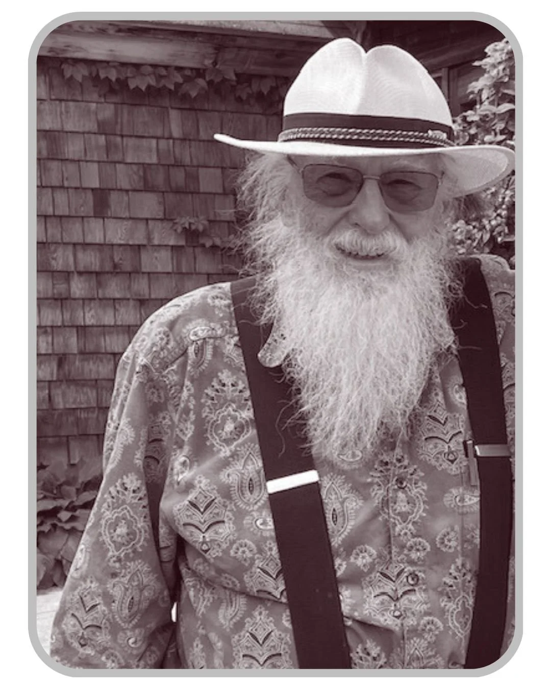 An elderly man with a long white beard, wearing sunglasses, a wide-brimmed hat, a patterned shirt, and suspenders, smiling outdoors in front of a wooden fence and foliage.