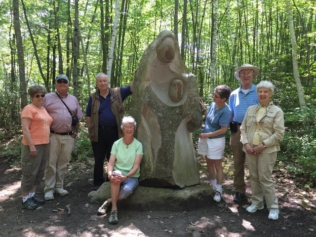 Seven people standing and sitting in a wooded area around a large stone sculpture with a face carved into it.