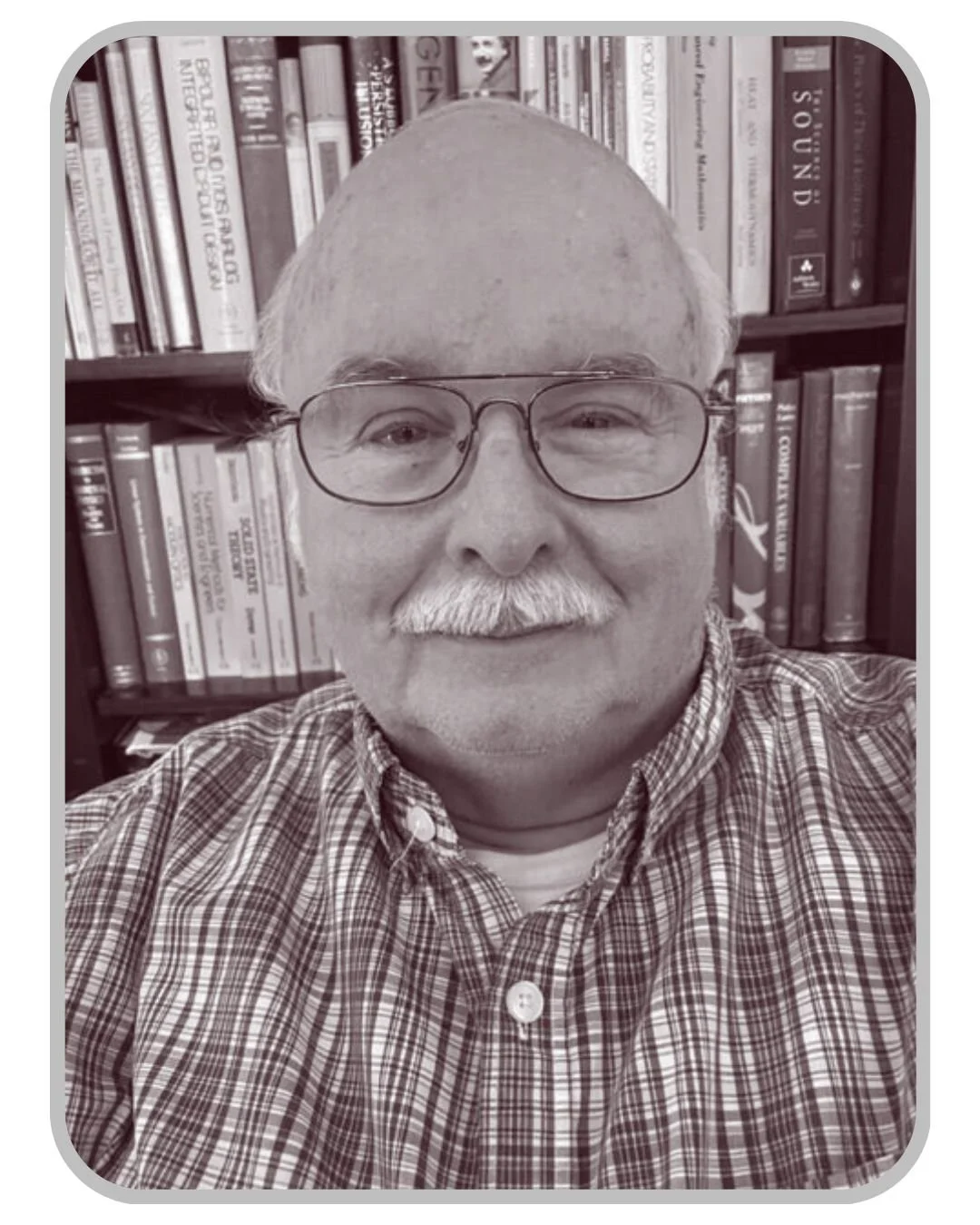A black-and-white selfie of an older man with glasses, a mustache, and a plaid shirt, standing in front of bookshelves filled with books.