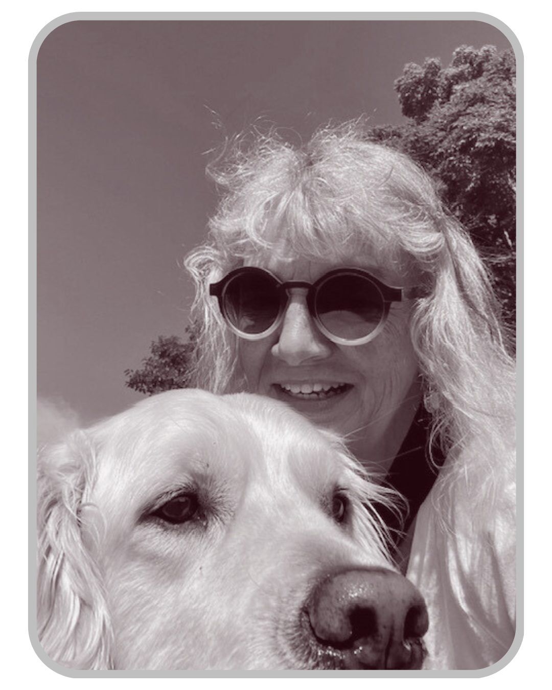 A woman with curly hair and sunglasses smiling next to a large light-colored dog outdoors on a sunny day.