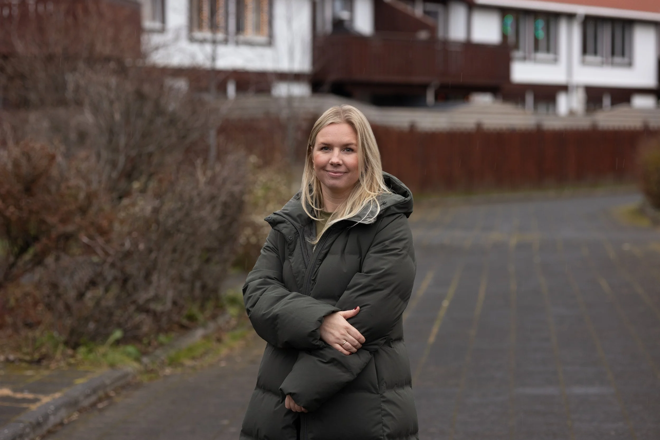 A woman with blonde hair wearing a dark green puffy coat, standing outdoors on a cloudy day in a parking lot with bushes and residential buildings in the background.