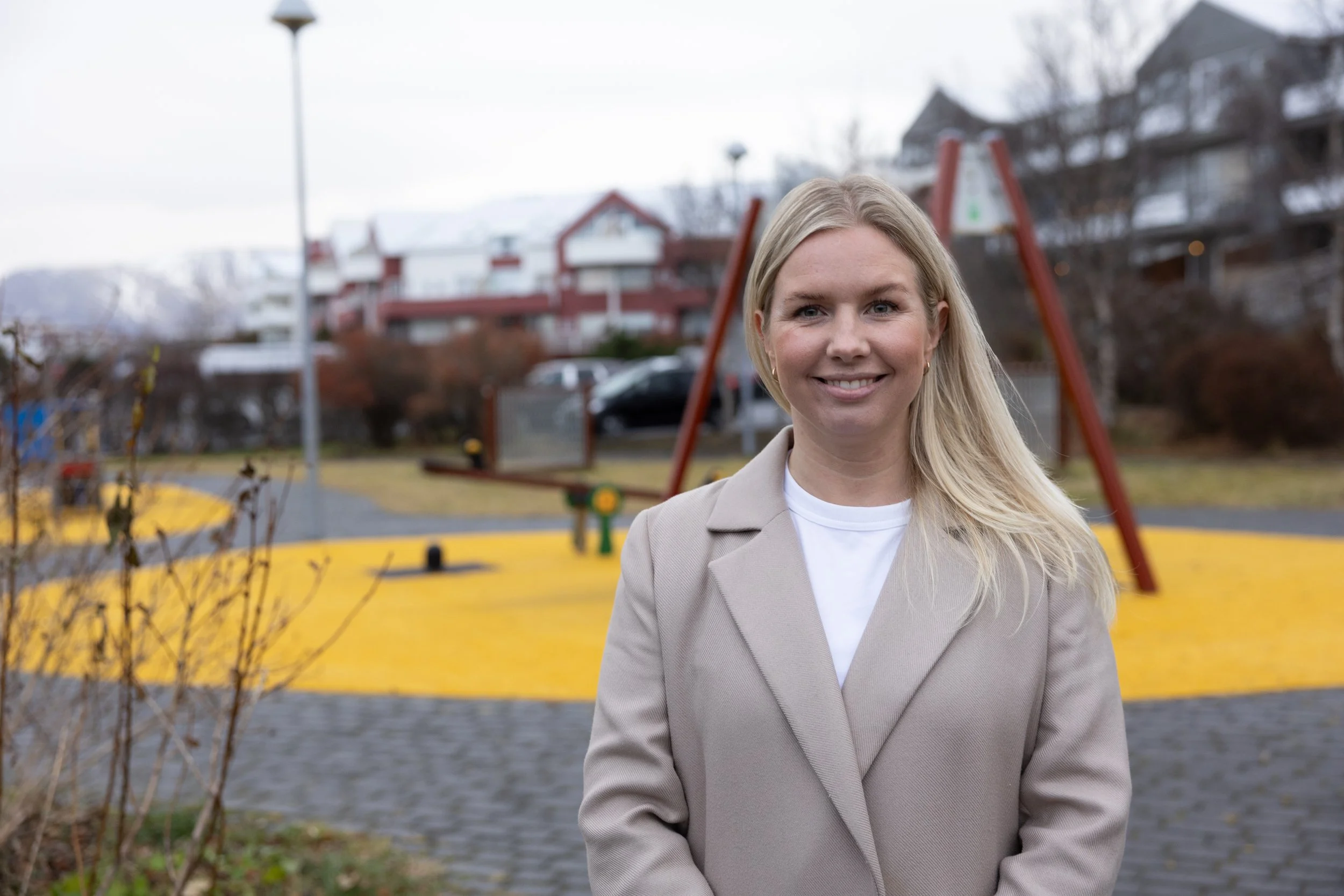 A woman with long blonde hair smiling outdoors near a playground with yellow surfaces, red swing set, and residential buildings in the background.