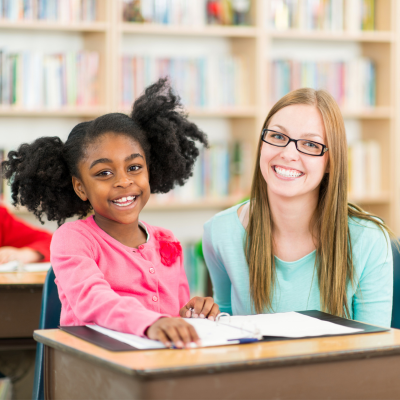A young girl and a woman smiling at a desk in a library, with bookshelves in the background.