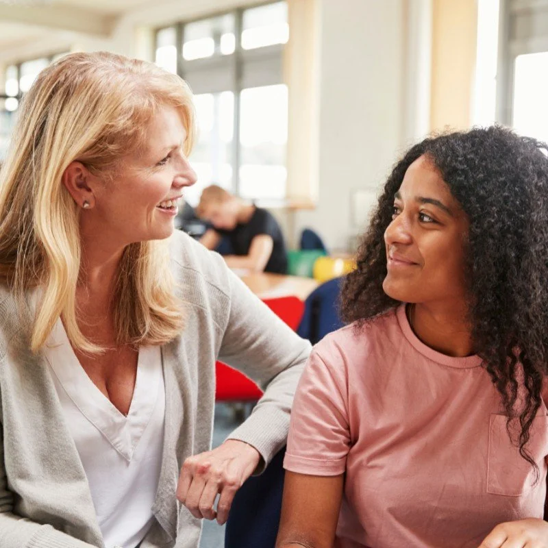 A woman with blonde hair, wearing a white shirt and gray cardigan, smiles and leans towards a young girl with curly black hair, wearing a pink T-shirt, in a bright room with large windows and other people in the background.