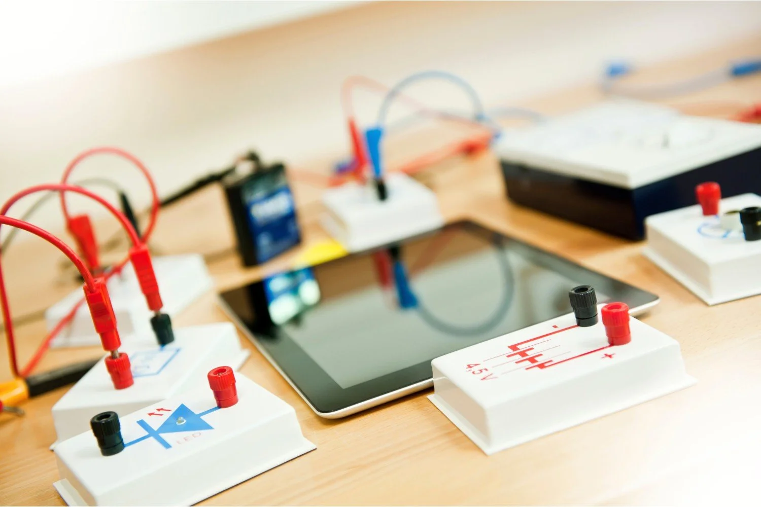 Educational electronic circuit experiments on a wooden table, including a tablet and various module boards with wires connected.