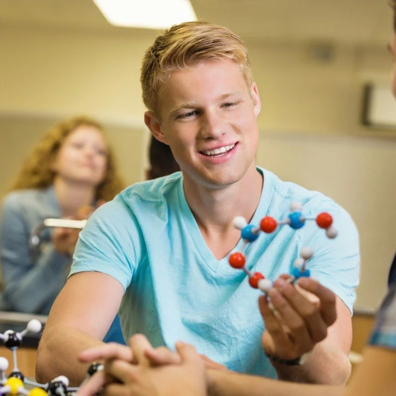A smiling young man in a light blue t-shirt holding a molecular model in a classroom setting, with a slightly out-of-focus girl in the background also holding a molecular model.