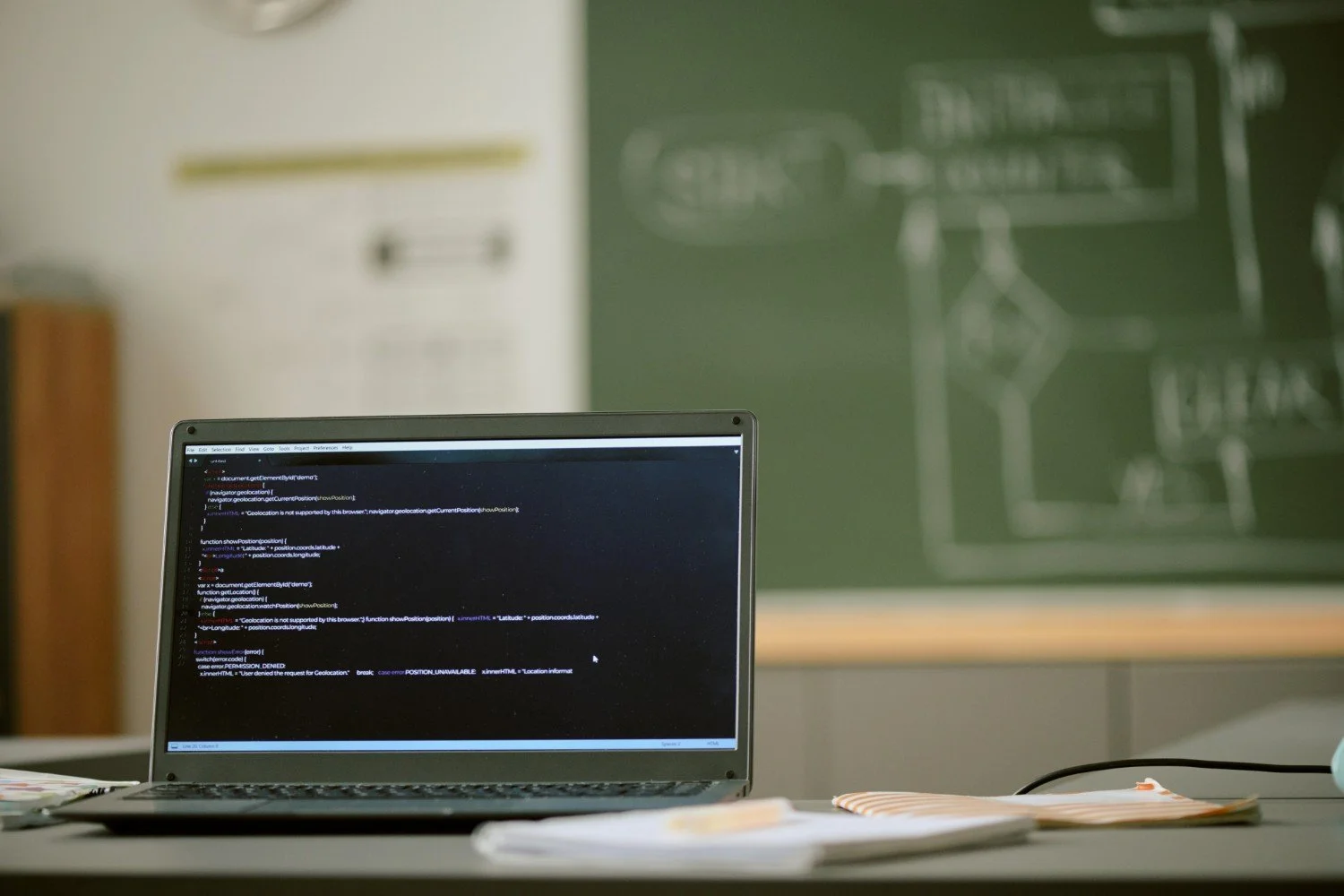 A laptop on a desk displaying programming code, with a blurred chalkboard or blackboard in the background.