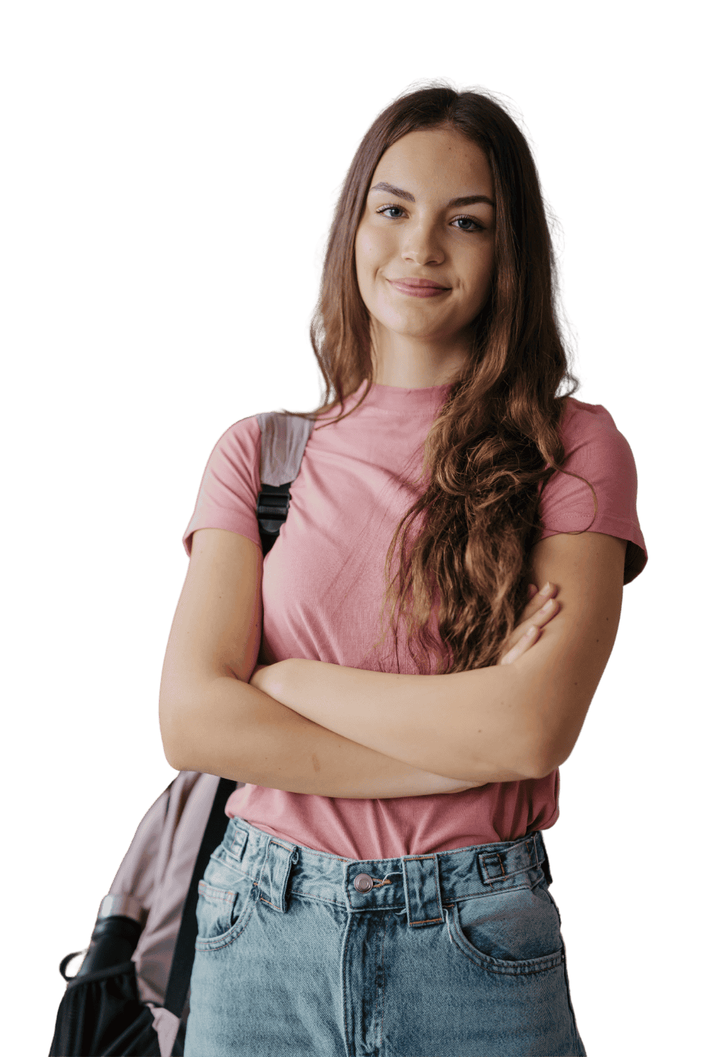 A young woman with long wavy brown hair, wearing a pink t-shirt and high-waisted denim shorts, standing with her arms crossed and smiling. She has a backpack on her shoulder.