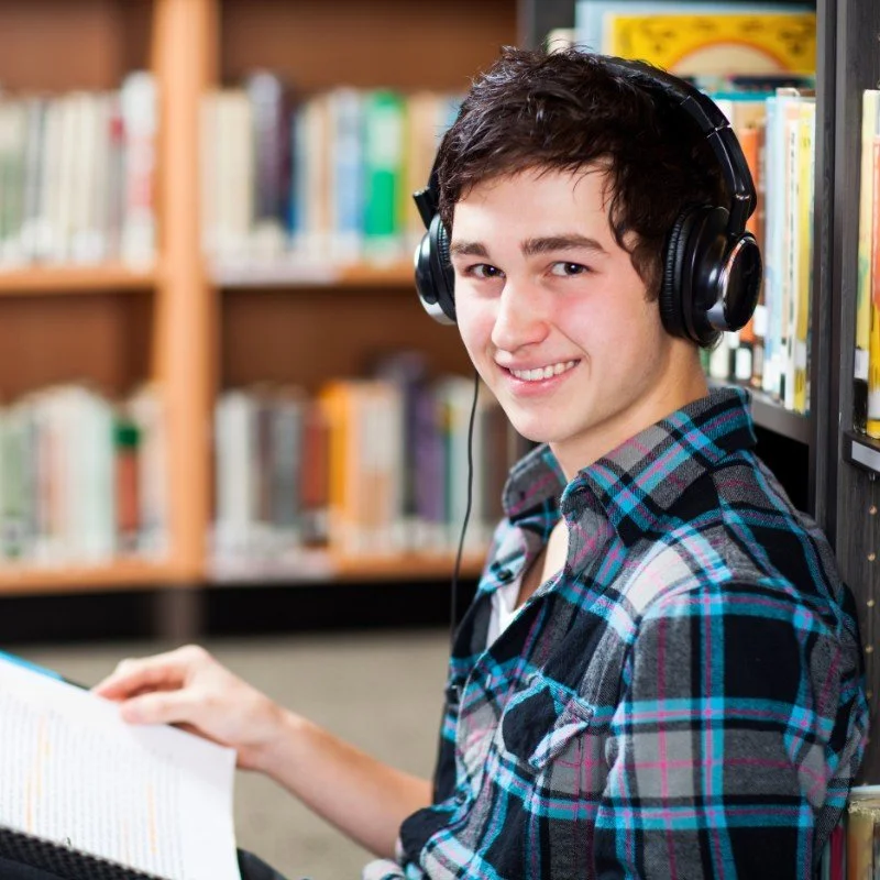 Smiling young man wearing headphones in a library, holding an open book.