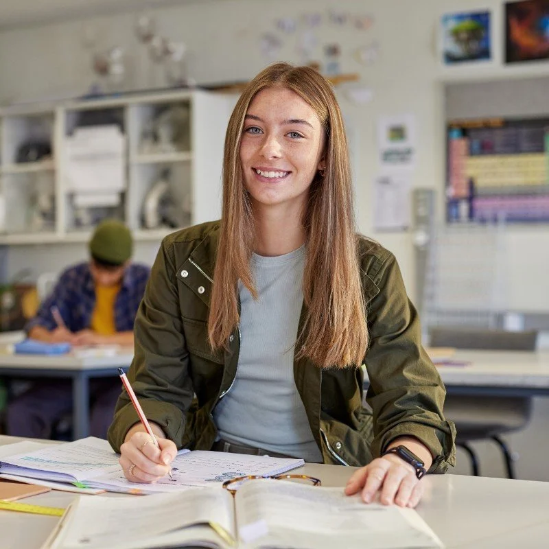 A young woman with long reddish-brown hair, sitting at a desk in a classroom, smiling at the camera, with school supplies and an open book in front of her. In the background, a person wearing a green beanie is writing at another desk.