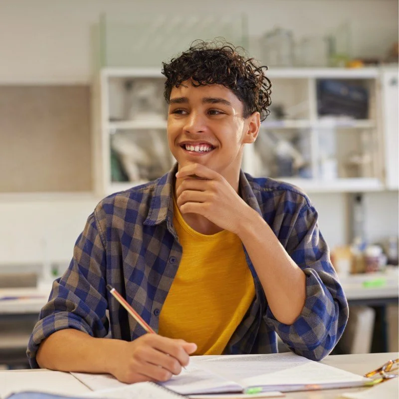 A young person with curly hair sitting at a desk with a pencil in hand, smiling and resting their chin on their hand, in a classroom or study environment.