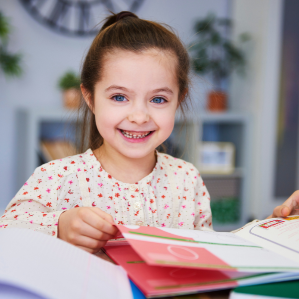 A young girl with blue eyes and brown hair tied back, smiling and looking at the camera while sitting at a desk with open colored notebooks.