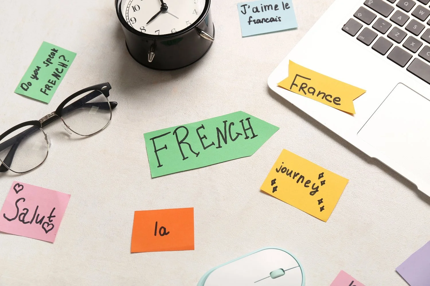 A cluttered desk with colorful sticky notes labeled 'French,' 'J'aime le français,' 'France,' 'journey,' 'Salute,' and 'la.' There are glasses, a black alarm clock, a laptop, and a white mouse on the desk.