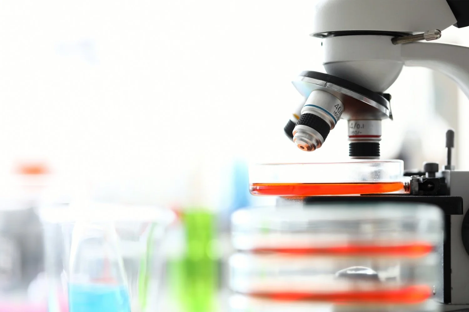 Microscope focusing on a petri dish with a red liquid in a laboratory setting.