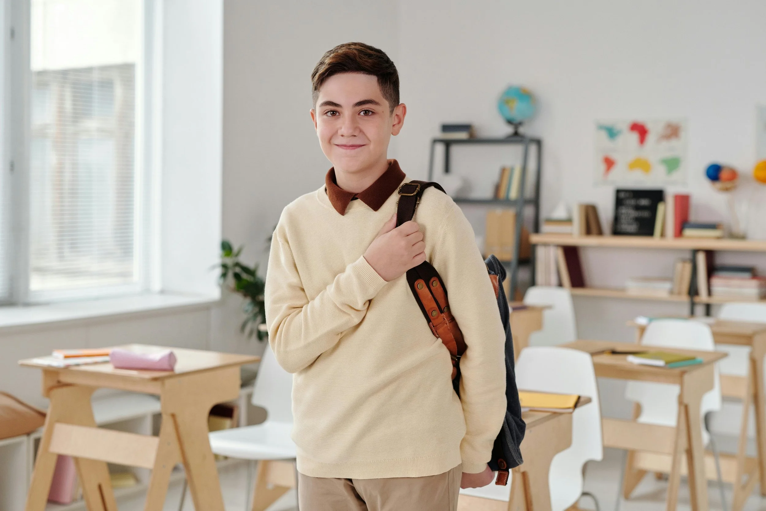 A young boy with short brown hair and light skin standing in a brightly lit classroom, carrying a backpack on his shoulder. He is wearing a beige sweater over a brown collared shirt and has a slight smile.