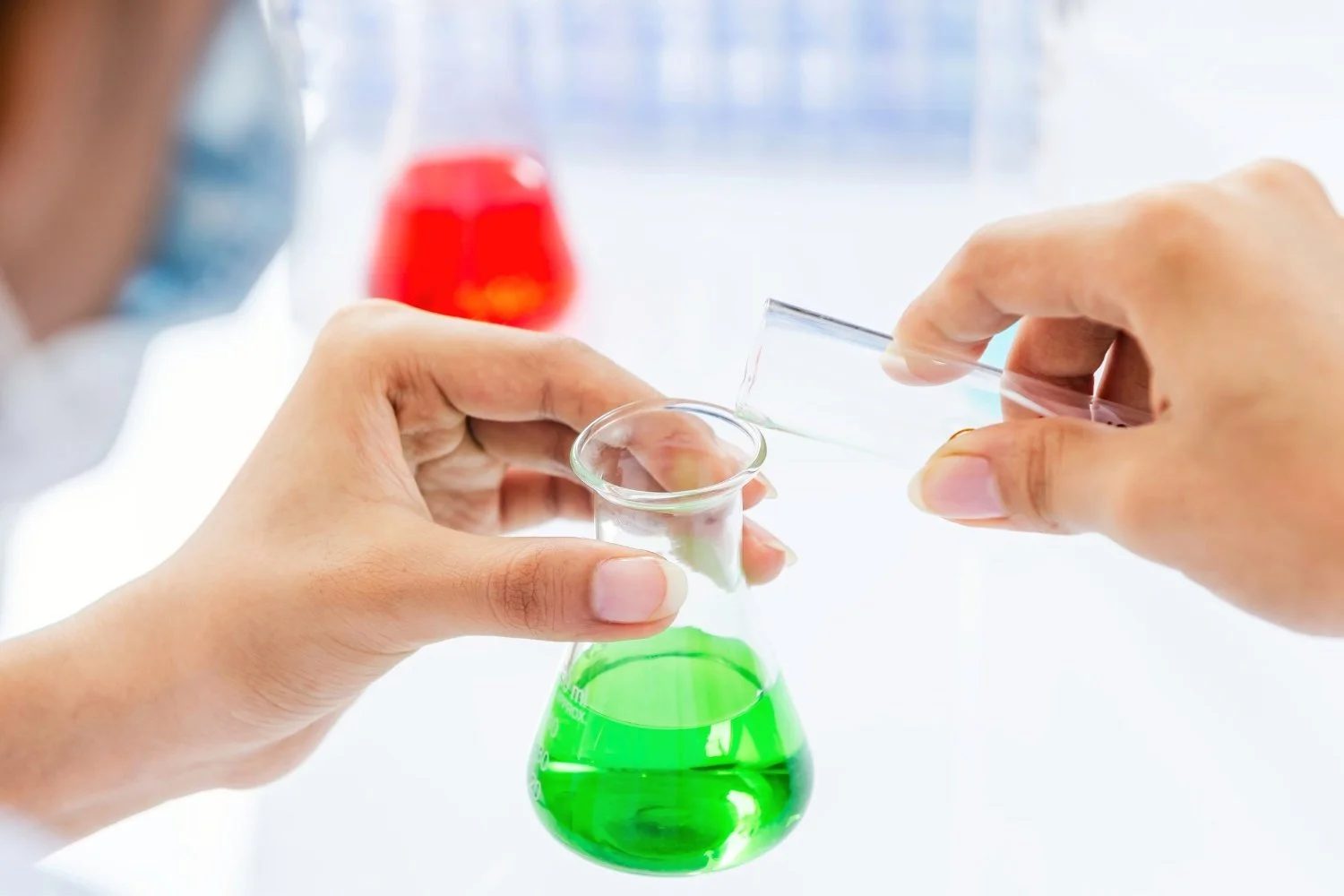 Person pouring a clear liquid into a test tube from a glass container, with a beaker of green liquid in front and a blurred red container in the background.