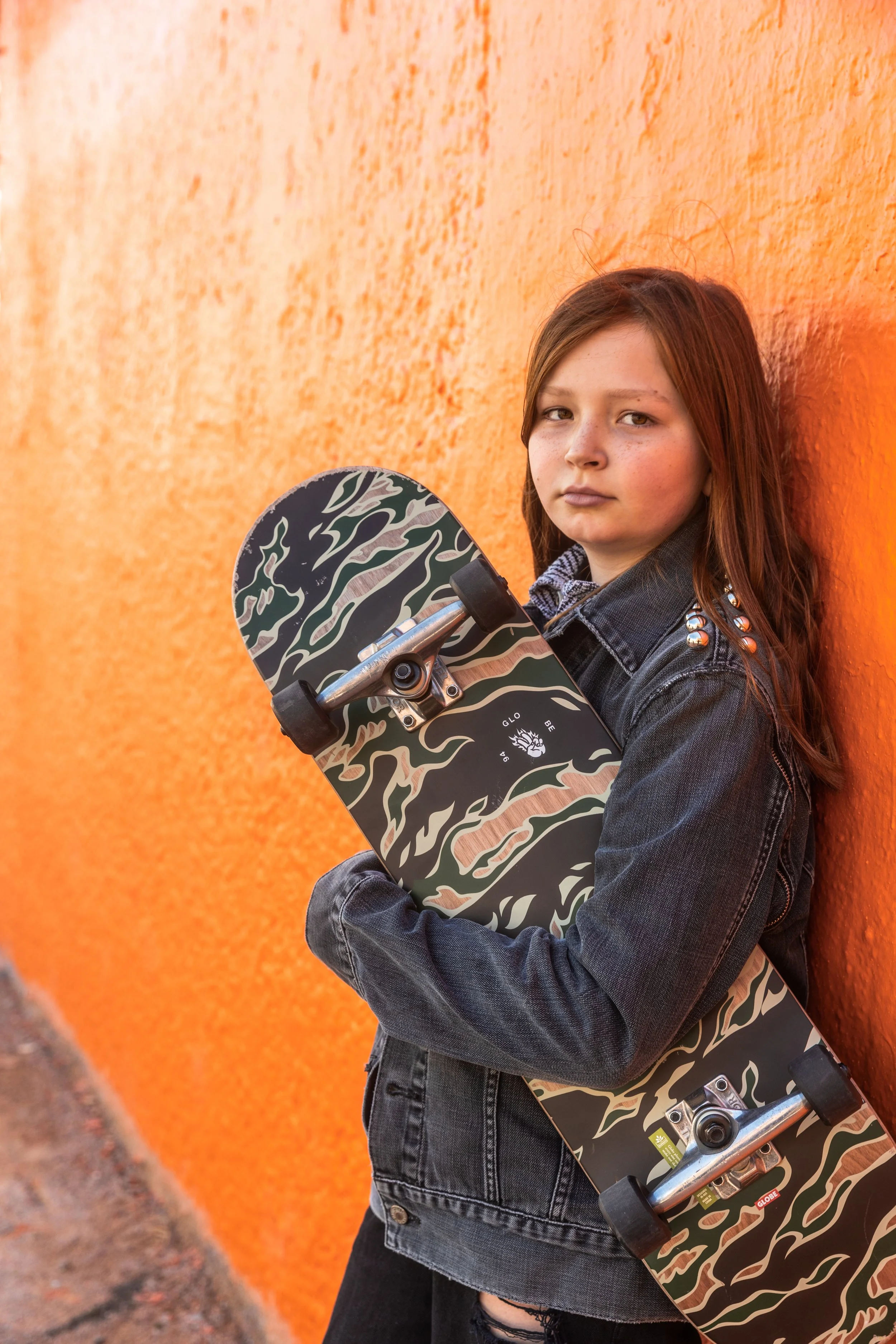 Jeune fille aux cheveux bruns portant une veste en jean avec des clous, tenant un skateboard à camouflage contre un mur orange.