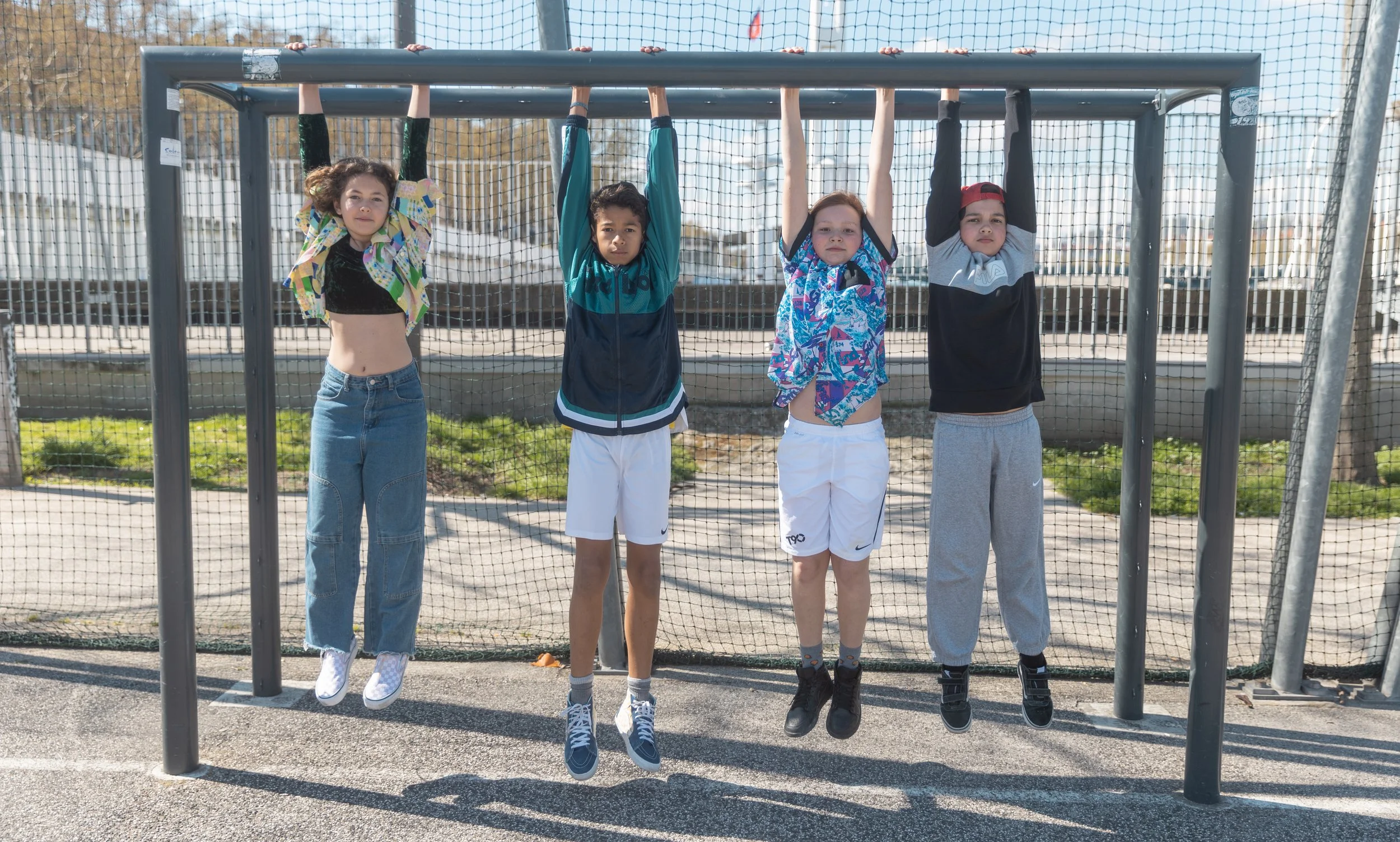 Quatre enfants suspendus à la barre d'une cage de but de basketball dehors, avec un filet derrière eux.
